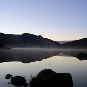 Fog drifting over a mirror-calm lake with dark stones in the shallows