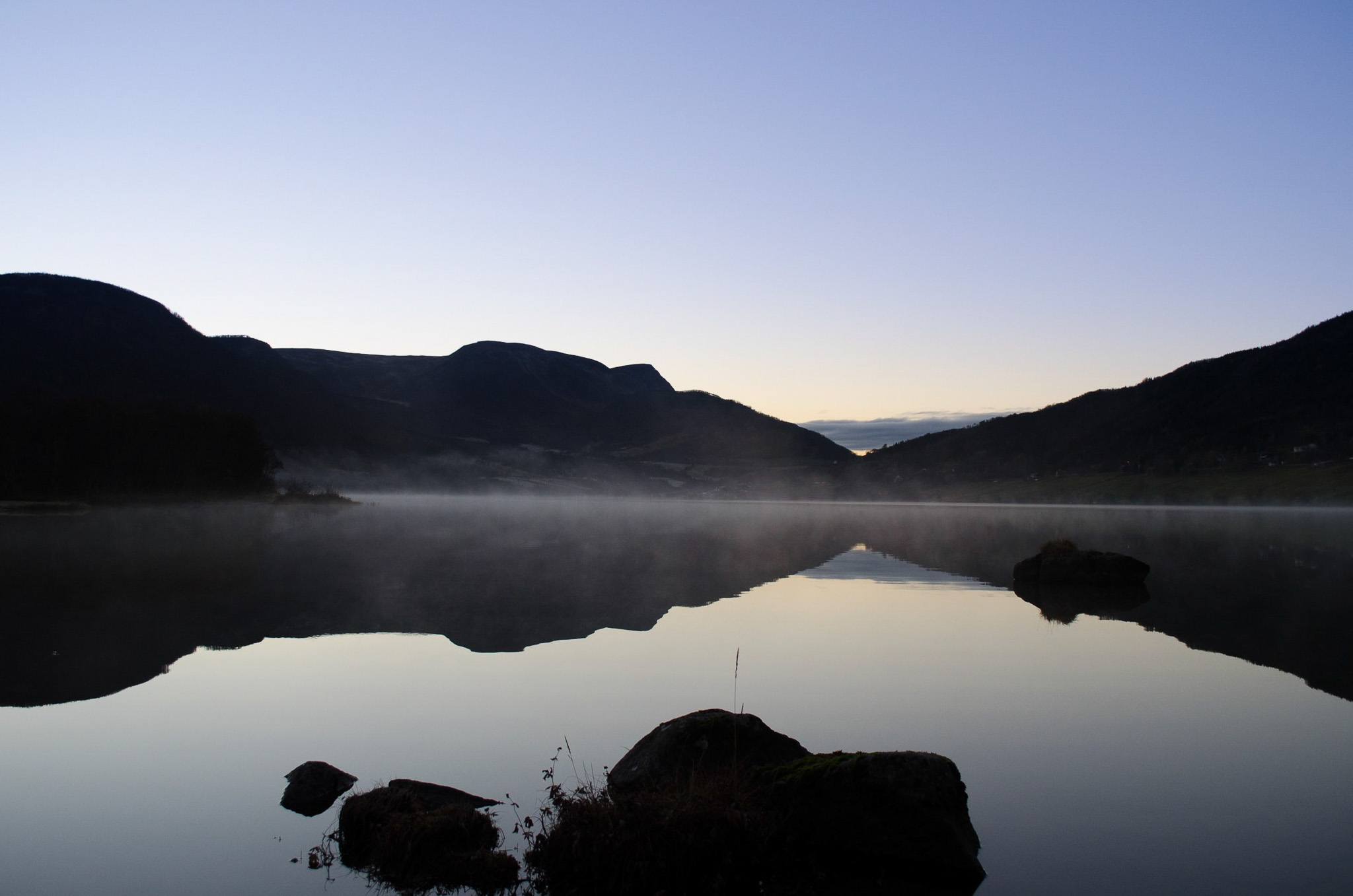 Fog drifting over a mirror-calm lake with dark stones in the shallows