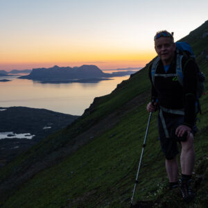 A hiker with poles climbing a green slope above a sunset fjord
